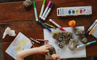 “Child’s hands arranging decorated stones on paper, surrounded by markers, crayons, paint, and art supplies on a wooden table.