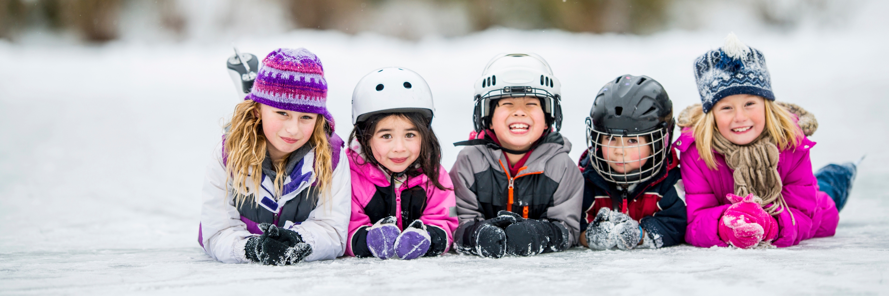 Group of children wearing winter helmets and hats lying on the ice and smiling outdoors.